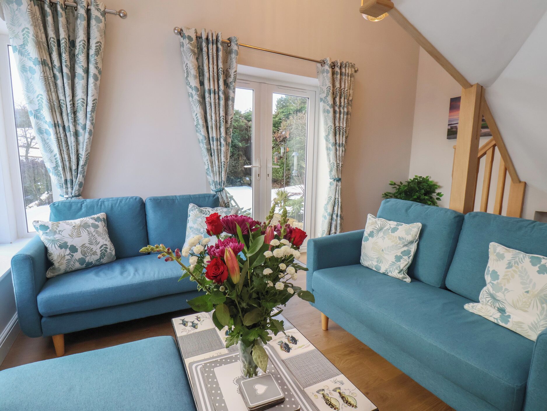 Een woonkamer met een blauwe bank, bloemkussens en een vaas met bloemen.