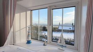 A room with a large window overlooking a snowy landscape, a bed with a white bedspread, and nautical-themed decorations on the windowsill. - Cheyne Cottage (Whitby)