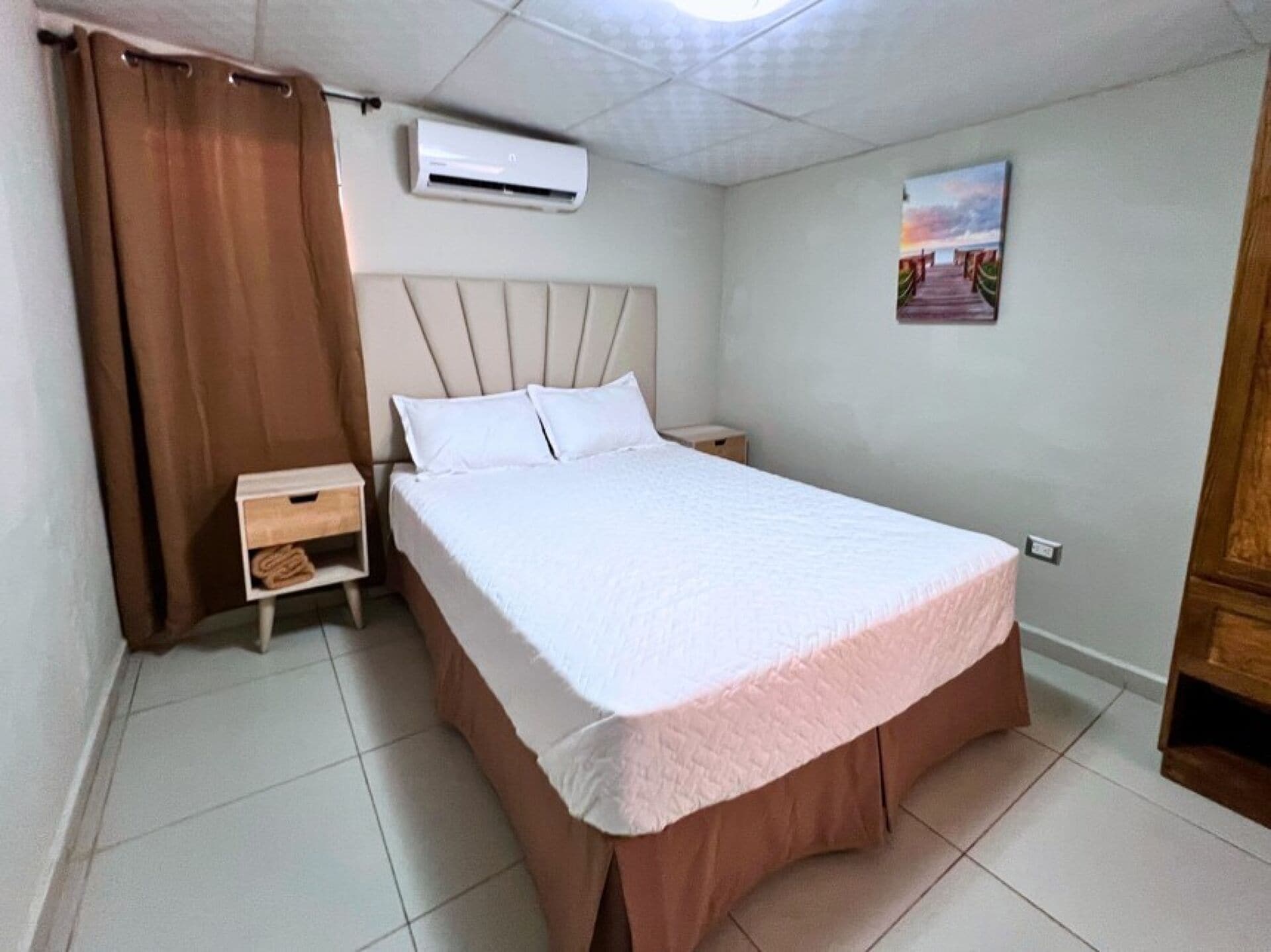 Una habitación de hotel con cama, mesita de noche, unidad de aire acondicionado, un cuadro en la pared y cortina.