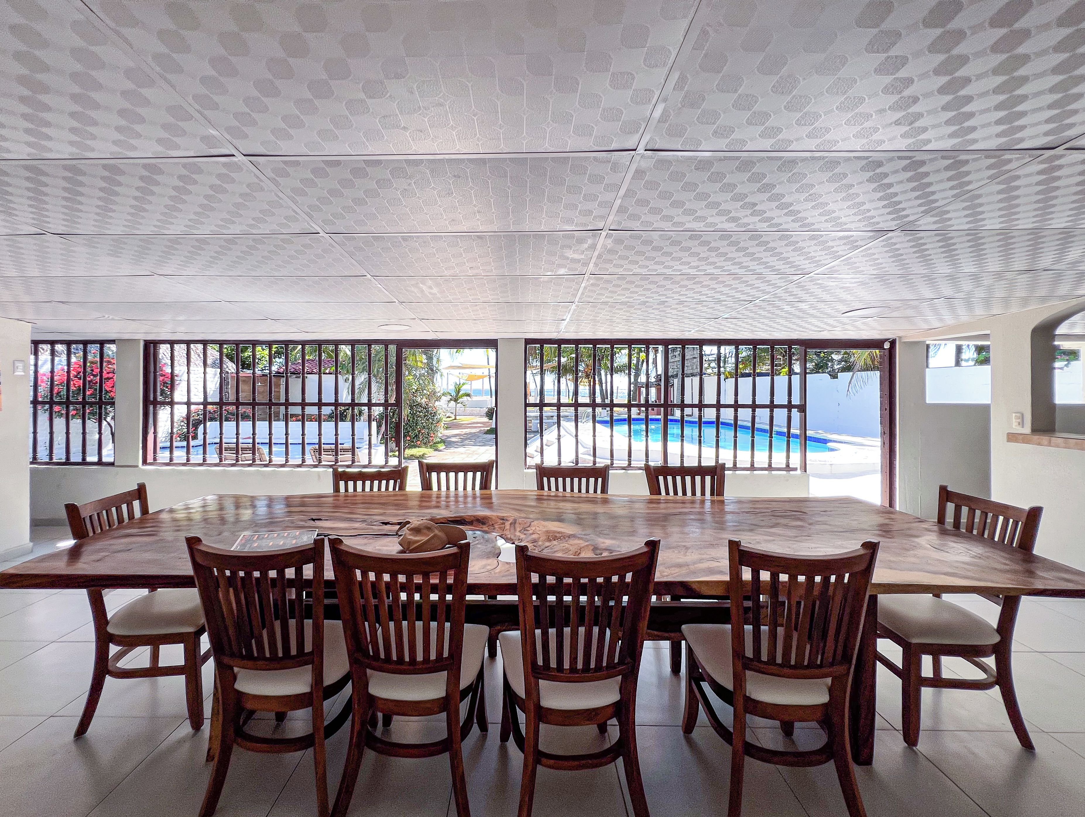 A dining area with a large table and chairs, a view of a swimming pool through barred windows, and a ceiling with a geometric pattern.