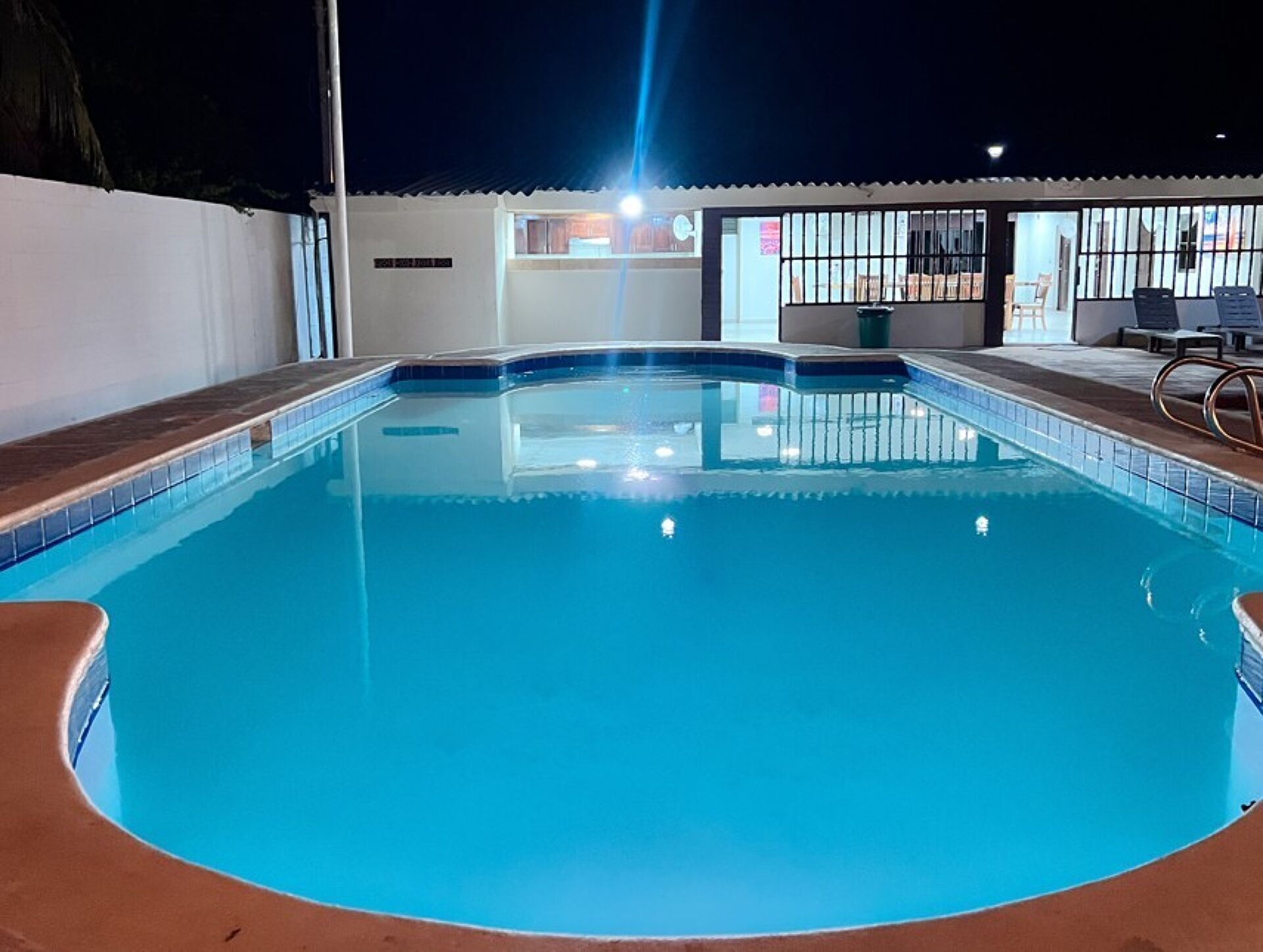 Una piscina con borde de azulejos, una zona cubierta con asientos y un edificio con un cartel al fondo.