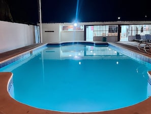 A swimming pool with a tiled edge, a covered area with seating, and a building with a sign in the background. - Brisas Del Surf - Conchalio Beach (La Libertad)