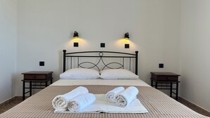 Ein ordentlich bezogenes Bett mit zwei Kissen, einem aufgerollten Handtuch und einem gefalteten Handtuch auf einer gemusterten Bettdecke. An beiden Seiten des Bettes stehen jeweils Nachttische mit Lampen.