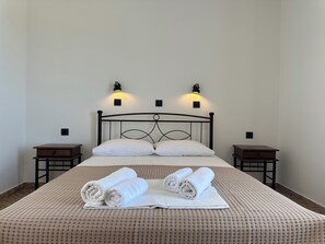 Ein ordentlich bezogenes Bett mit zwei Kissen, einem aufgerollten Handtuch und einem gefalteten Handtuch auf einer gemusterten Bettdecke. An beiden Seiten des Bettes stehen jeweils Nachttische mit Lampen.