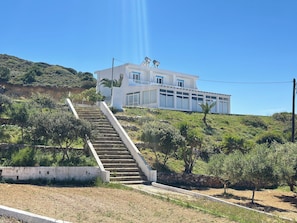 A white building with a staircase leading up to it, surrounded by greenery and hills. - Maistros SeaView Suite (Kos)