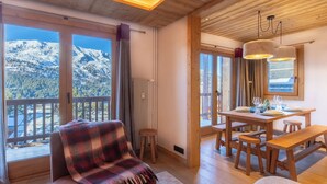 Una habitación acogedora con techo de madera, una manta escocesa sobre una silla, una mesa de comedor con platos y vasos, y vistas a montañas nevadas a través de ventanas amplias.