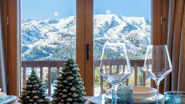 Una mesa de comedor con dos copas de vino y un pequeño árbol de Navidad, con el paisaje nevado de una montaña al fondo visible a través de amplios ventanales.