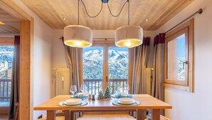 Una mesa de comedor de madera con dos lámparas colgantes, un pequeño árbol de Navidad y vistas a montañas nevadas a través de un amplio ventanal.