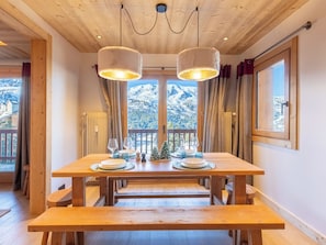 Ein Holztisch mit zwei hängenden Lampen, eine kleine Weihnachtstanne und Blick auf schneebedeckte Berge durch ein großes Fenster.