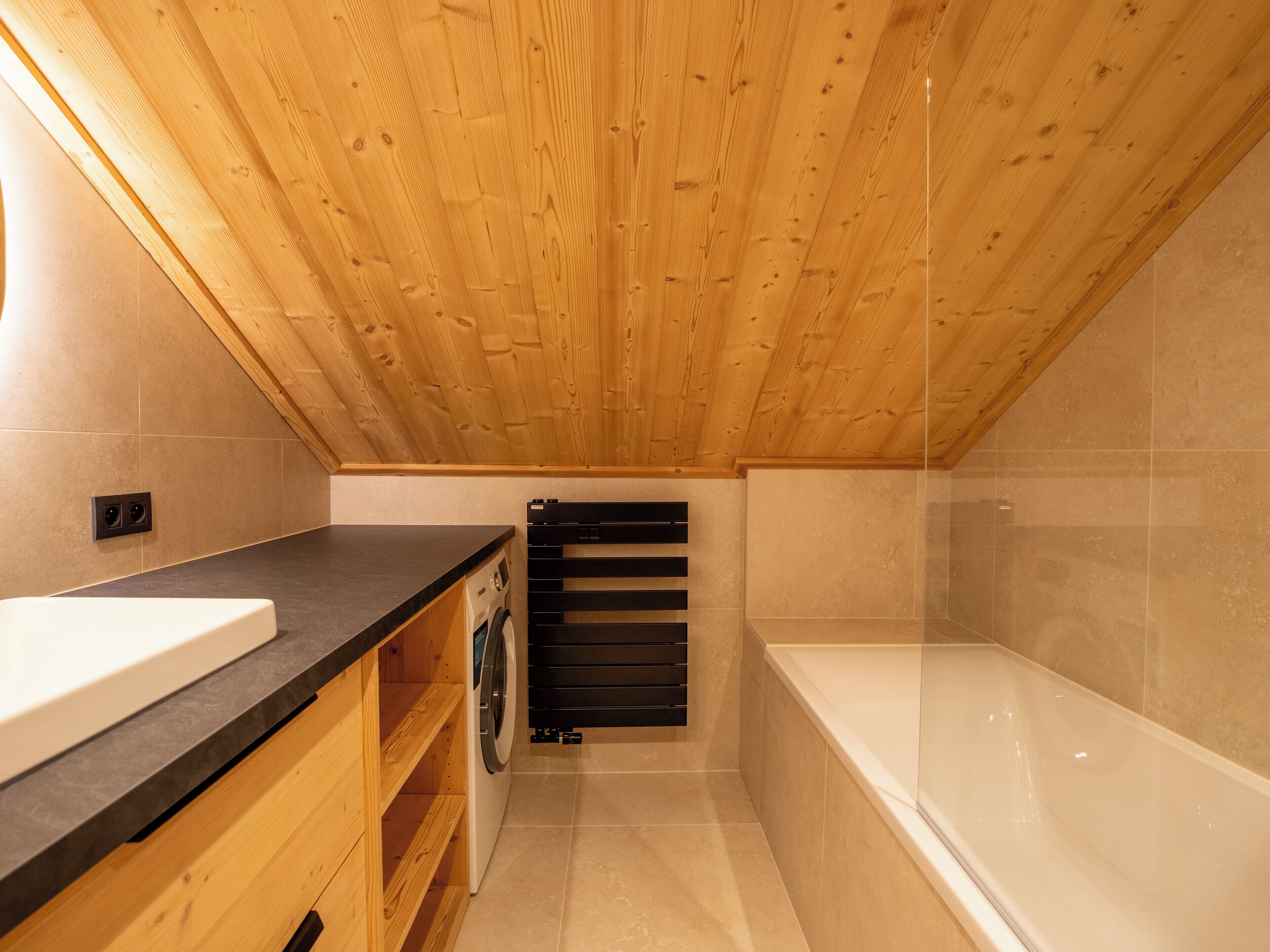 A small, modern bathroom with a wooden ceiling, a bathtub, a washing machine, and a sink.