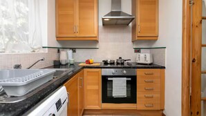 A compact kitchen with wooden cabinets, a stainless steel range hood, a built-in oven, and a small countertop with a kettle and a cutting board.