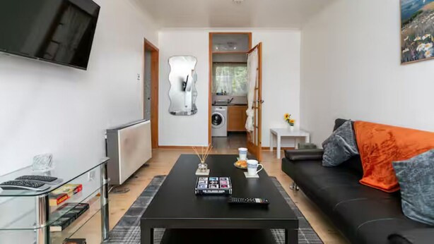 A modern living room with a black coffee table, a black sofa with orange and grey cushions, a glass coffee table with books and a remote control, a mounted TV, and a kitchen area with a washing machine.