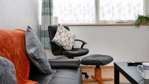 A modern living room with a black leather sofa, a black office chair with a floral cushion, a wooden coffee table, and a window with sheer curtains.