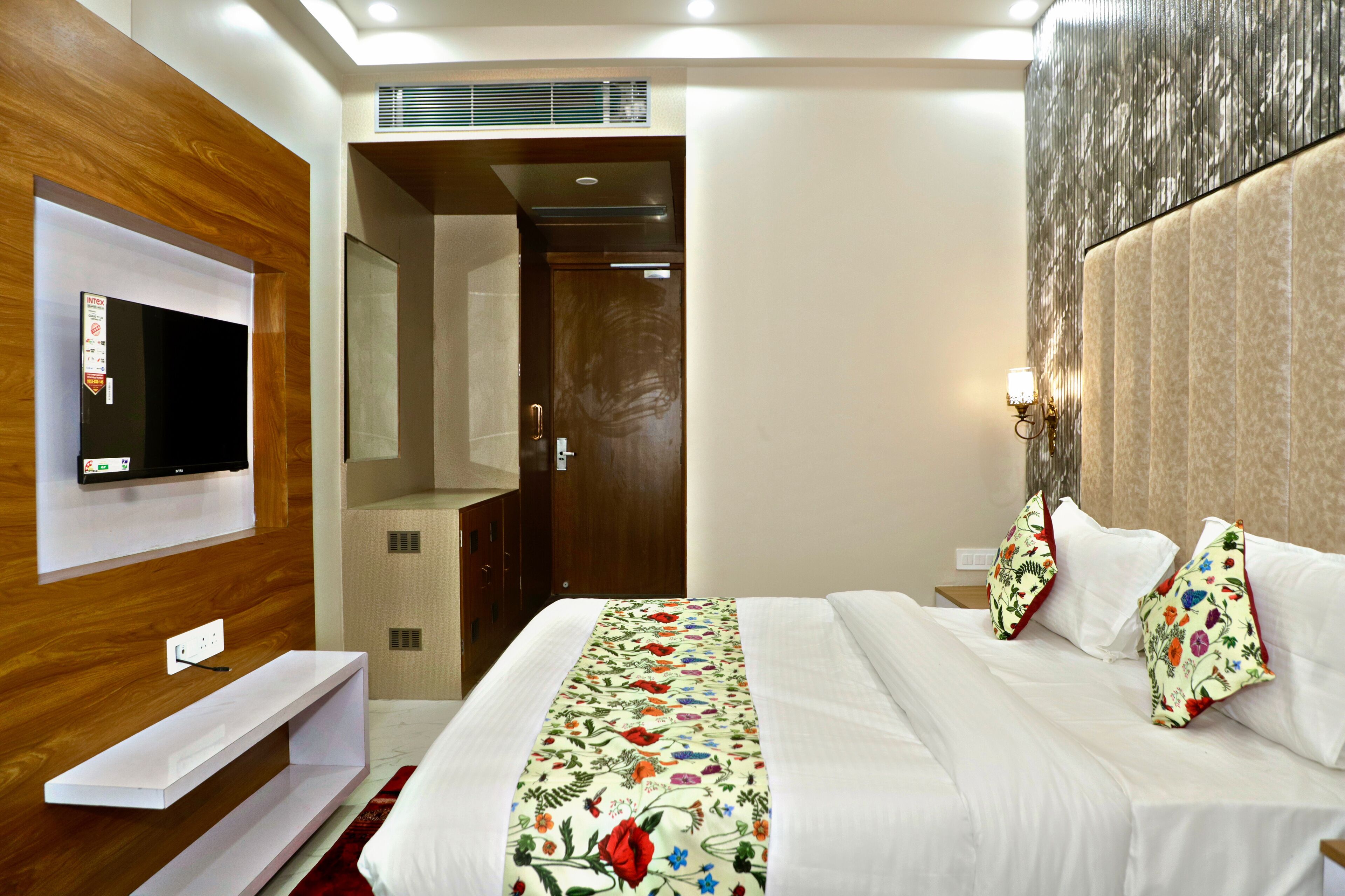 Habitación de hotel con cama, televisor montado en la pared, paneles de madera, una puerta y una mesita de noche con lámpara.
