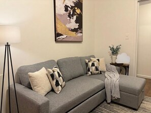 A grey sofa with patterned cushions, a floor lamp, a painting, a small table with a plant, and a white object on the table.