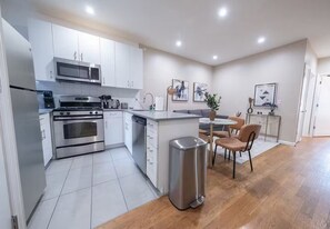 A modern kitchen with stainless steel appliances, a dining area with a table and chairs, and a trash bin. - Brand New Gorgeous 3bedroom 2blocks to CentralPark (New York)