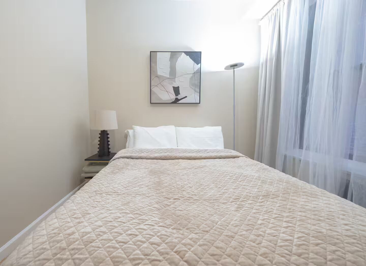 Ein Hotelzimmer mit Bett, Nachttisch mit Lampe, einem Bild an der Wand und durchsichtigen Vorhängen.
