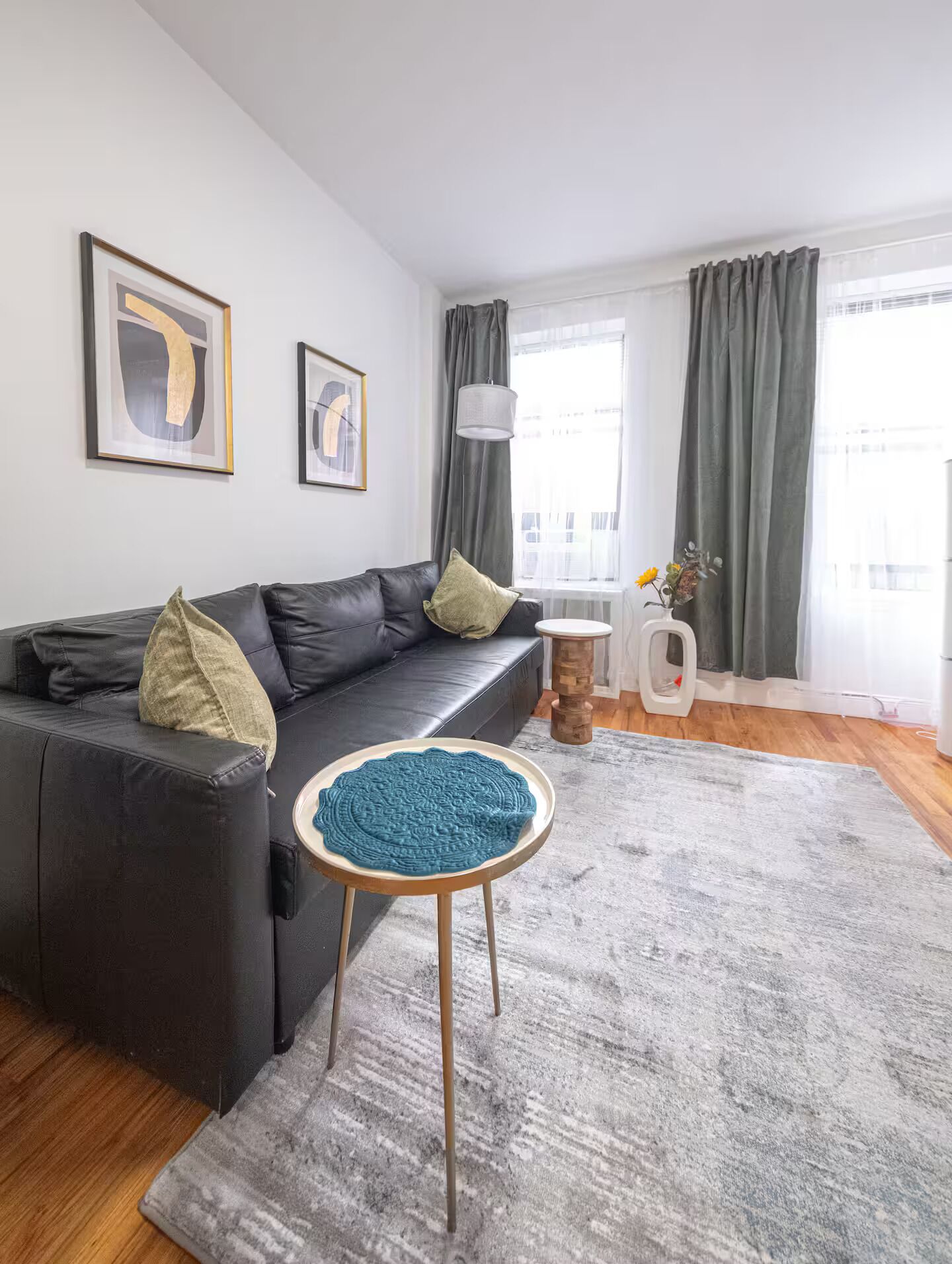 Ein modernes Wohnzimmer mit einem schwarzen Sofa, einem runden Holzschemel, einem grauen Teppich und zwei gerahmten Kunstwerken an der Wand.