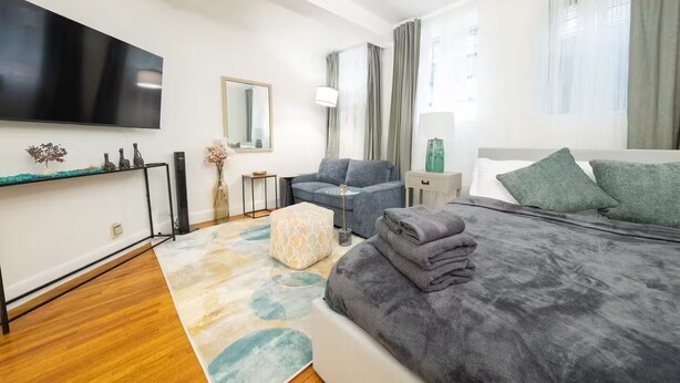 Moderni hotellihuone, jossa on sänky, sohva, televisio, pieni pöytä ja ikkuna, jossa on verhot.