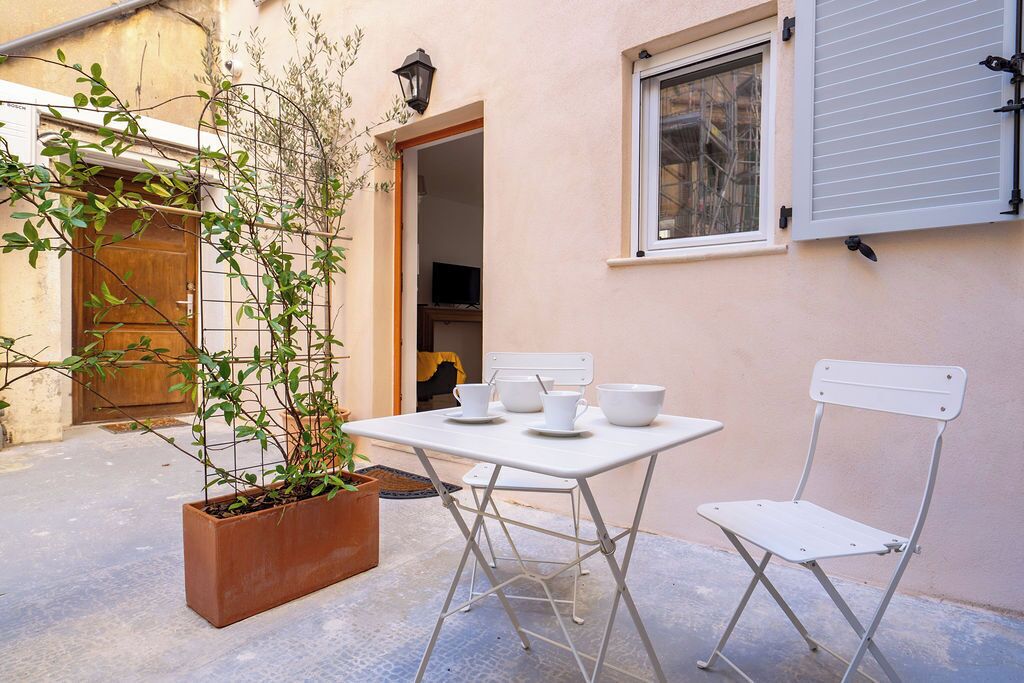 Un pequeño patio al aire libre con una mesa y dos sillas blancas, una planta en maceta y una puerta de madera.