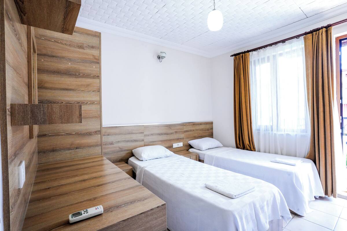 Ett hotellrum med två separata sängar, en sänggavel i trä, vita väggar och ett fönster med gardiner.