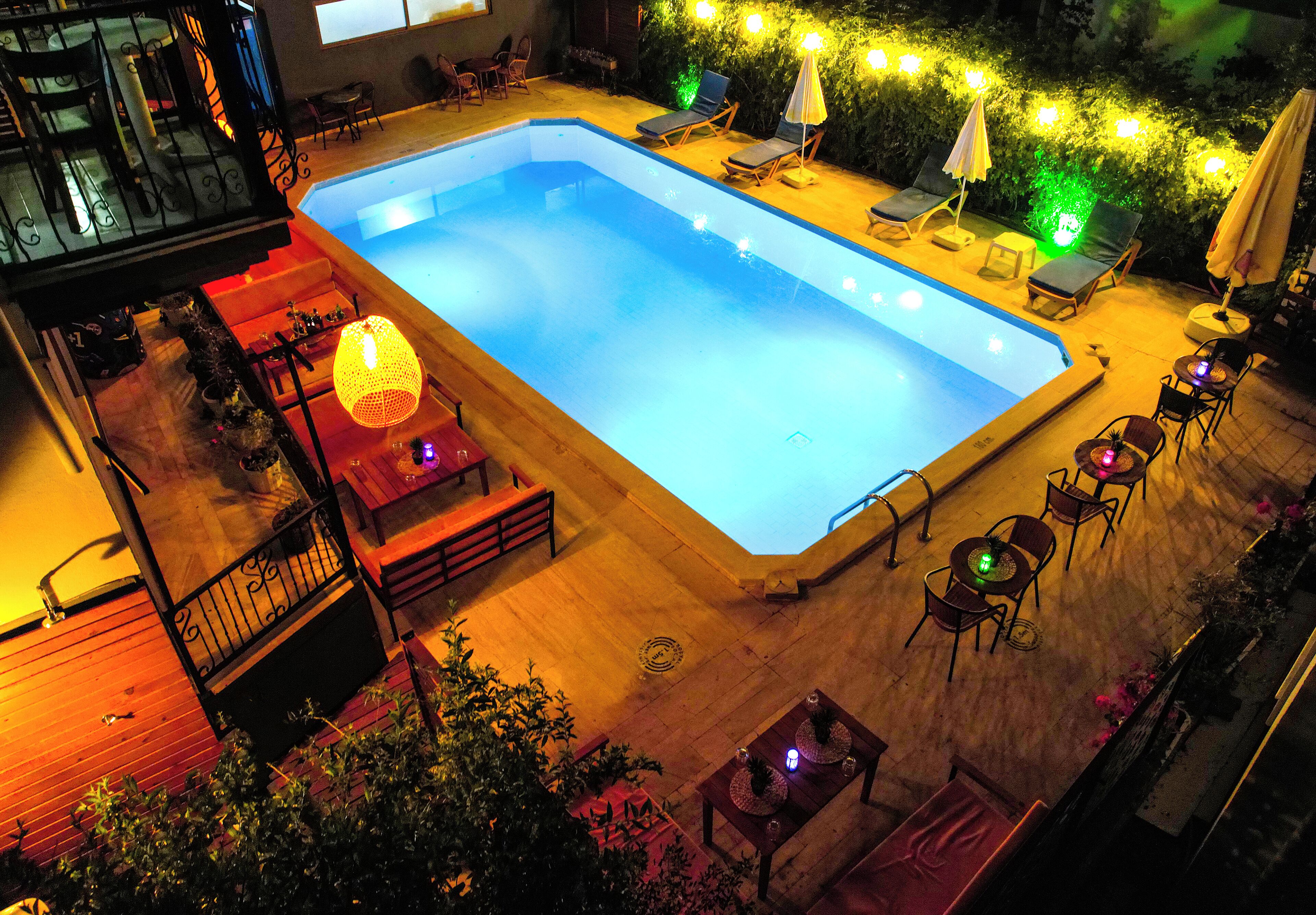 A rectangular swimming pool with lounge chairs and tables around it, surrounded by a wooden deck and outdoor seating area with string lights.