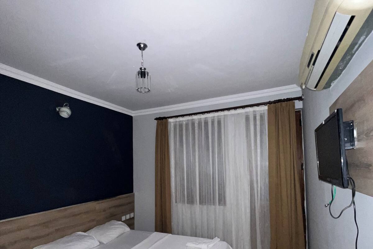 Ett hotellrum med en säng, en taklampa, en luftkonditioneringsanläggning, en tv och ett fönster med gardiner.