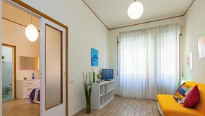 Un saló amb un sofà, una televisió sobre un moble, una finestra amb cortines i una porta que dona a una altra habitació.