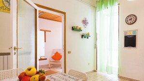Habitació amb taula de dinar posta, un bol de fruita, una cadira i un tapet de taula. Hi ha una porta que condueix a una altra habitació, una cadira amb coixí taronja i una estant amb objectes decoratius.