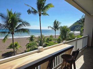A balcony with a view of a beach, palm trees, and a mountainous landscape. - Apartment by the Sea • Balcony with a view of the sunset - Access to the Crocs Resort - Direct beach access - (Jaco)