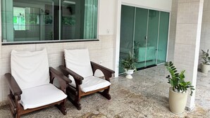A room with two wooden chairs with white cushions, a large window, a sliding glass door, and potted plants. - Quiet house - close to Vitória airport (Vitória)