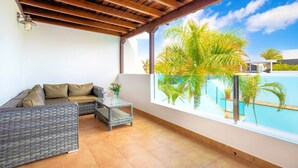A balcony with a wicker sofa, a small table with a vase, and a view of a swimming pool with palm trees. - Villa Puerto Calero Marina | Short Walk to Centre | Private Heated Pool (Lanzarote)