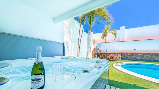 A hot tub with a bottle of champagne and glasses, a pool, and palm trees in the background. - Villa Puerto Calero Marina | Short Walk to Centre | Private Heated Pool (Lanzarote)