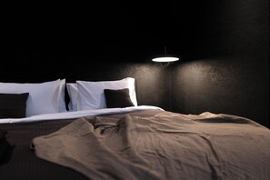 Una cama con ropa de cama blanca y marrón, una pared oscura y una única lámpara.