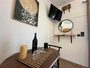 A room with a round wooden table, a bottle of wine, wine glasses, a wooden candle, a black bottle, a TV mounted on the wall, a mirror, and a shelf with decorative items. - Ostria SeaView Suite (Kos)