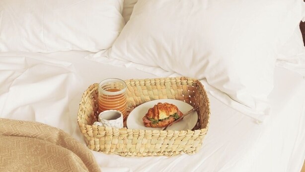 Una cama bien hecha con sábanas blancas, una bandeja de mimbre con un sándwich, una taza y una taza de café.