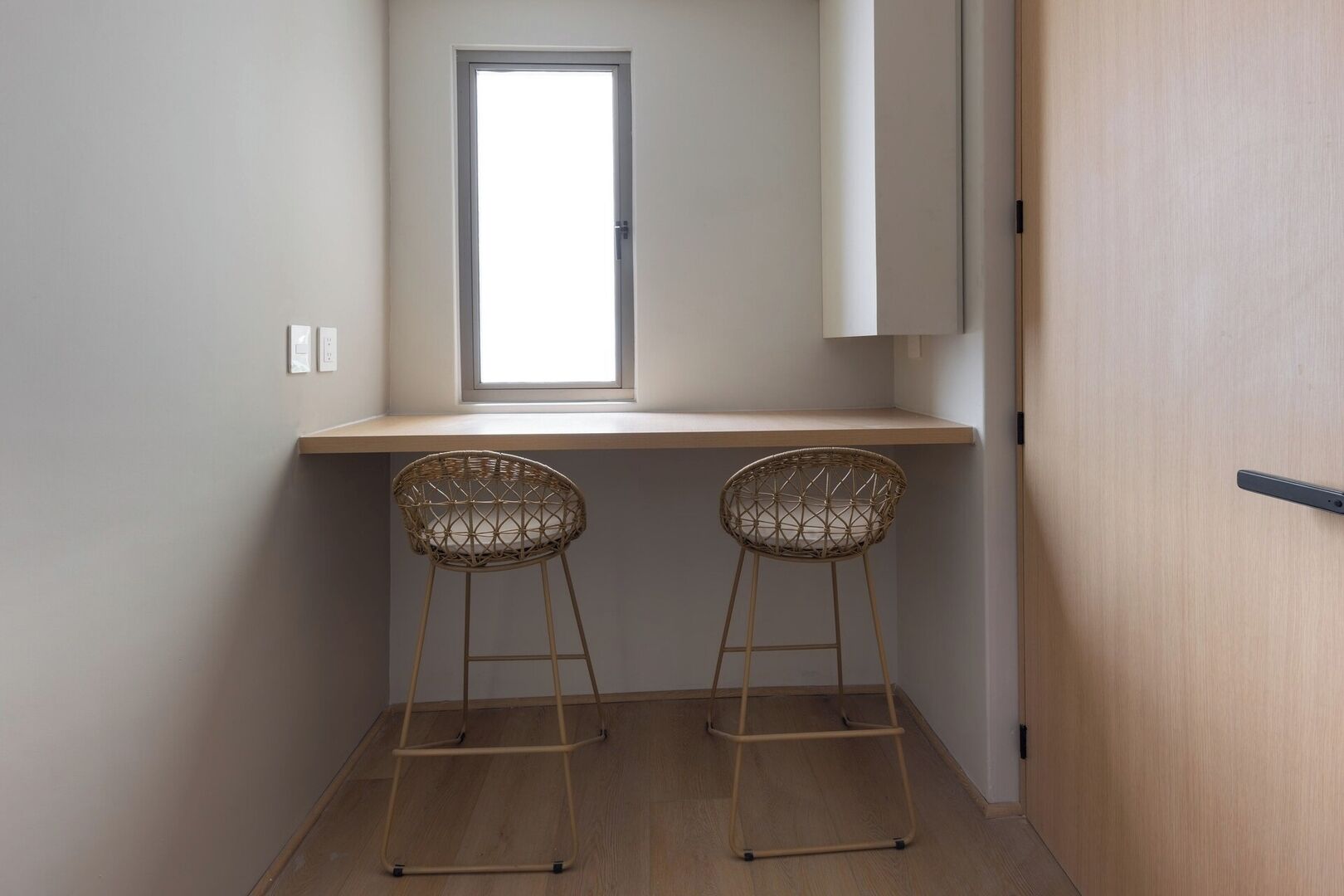 Une petite pièce avec une fenêtre, deux fauteuils en osier, un bureau en bois et une porte.