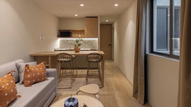 Ett kompakt bostadsutrymme med ett litet pentry, en soffa med mönstrade kuddar, ett litet runt bord och ett fönster med gardiner.