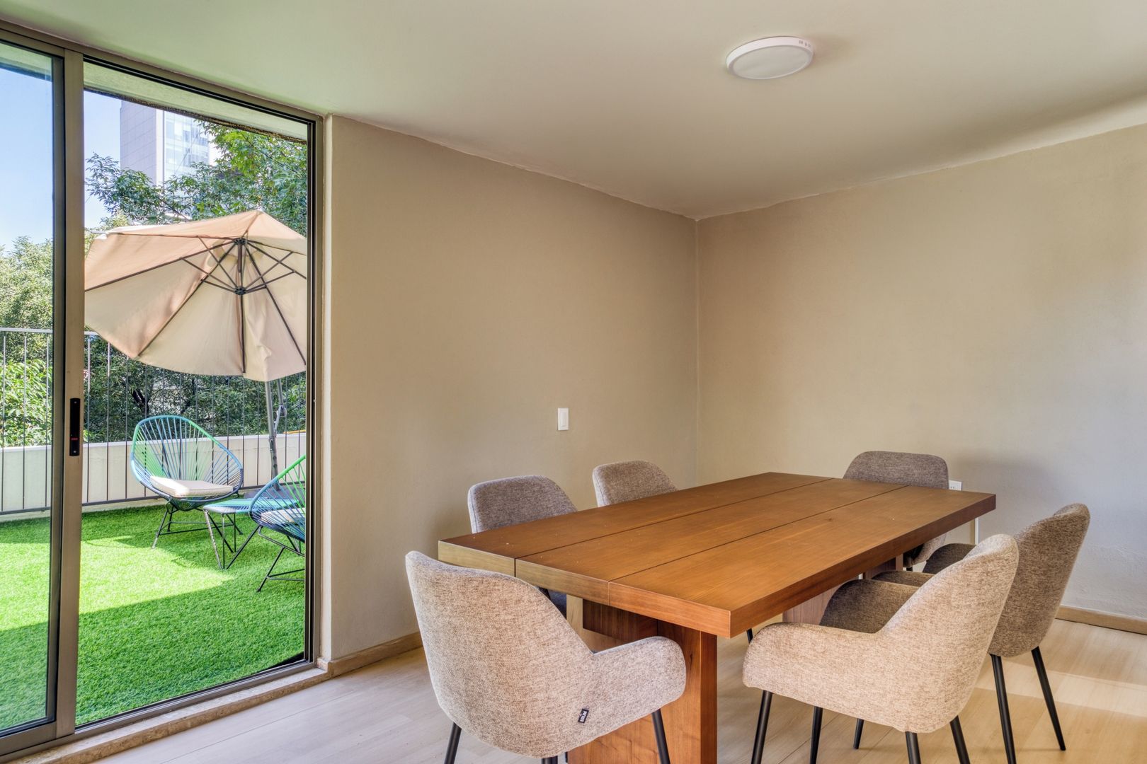 Un comedor con una mesa de madera y seis sillas, una puerta corrediza de vidrio que da a un jardín con sombrilla.