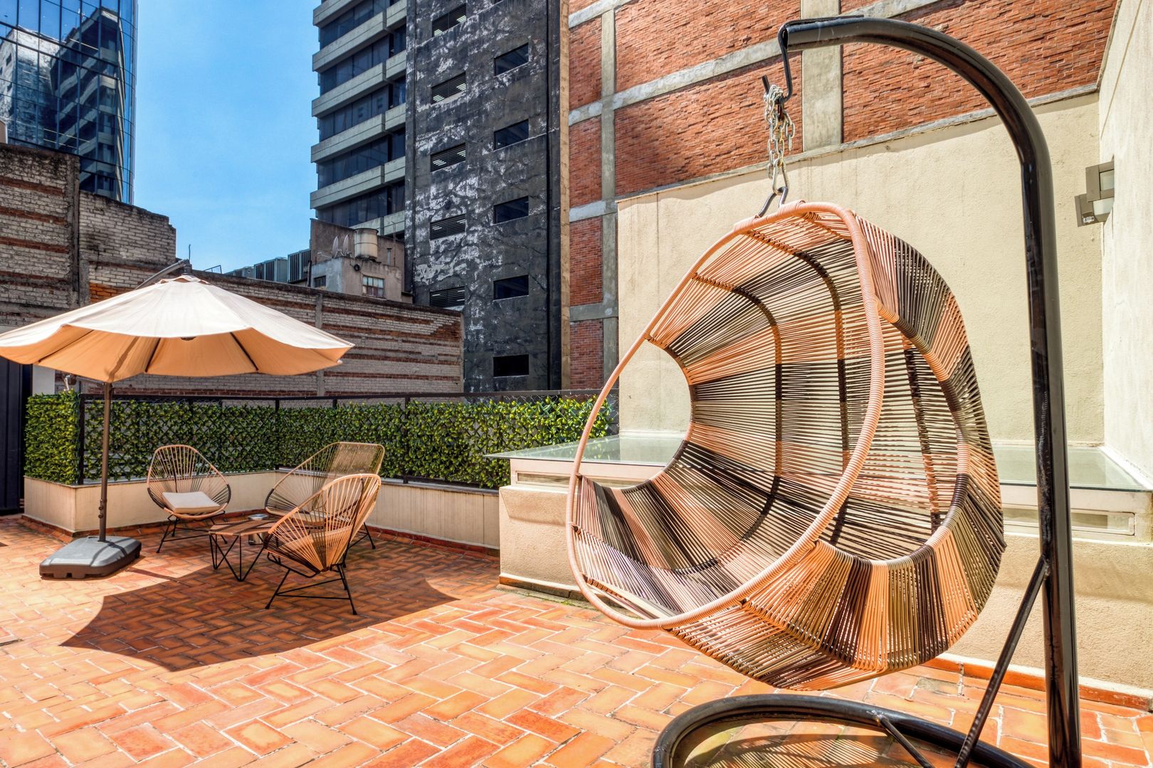 Une terrasse sur le toit, avec des chaises en osier, un fauteuil suspendu et une table placée sous un grand parasol.