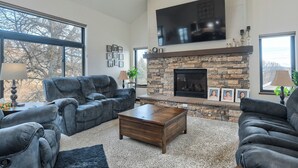 A living room with a stone fireplace, a flat-screen TV mounted above it, and a large window providing a view of trees outside. - Sturgis Motorcycle Rally Rental - 5 bedroom with garage (Spearfish)