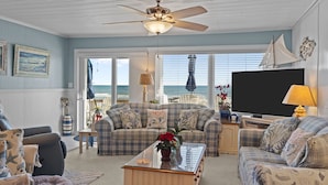 Ein Wohnzimmer mit großem Fenster zum Meer, Deckenventilator und Fernseher.