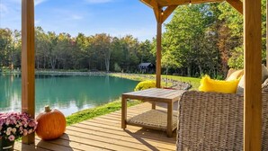 A wooden deck with a wicker sofa and a table, overlooking a calm lake and surrounded by trees.