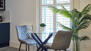 Un coin repas avec une table en verre et deux chaises, une plante en pot et une fenêtre avec des stores.