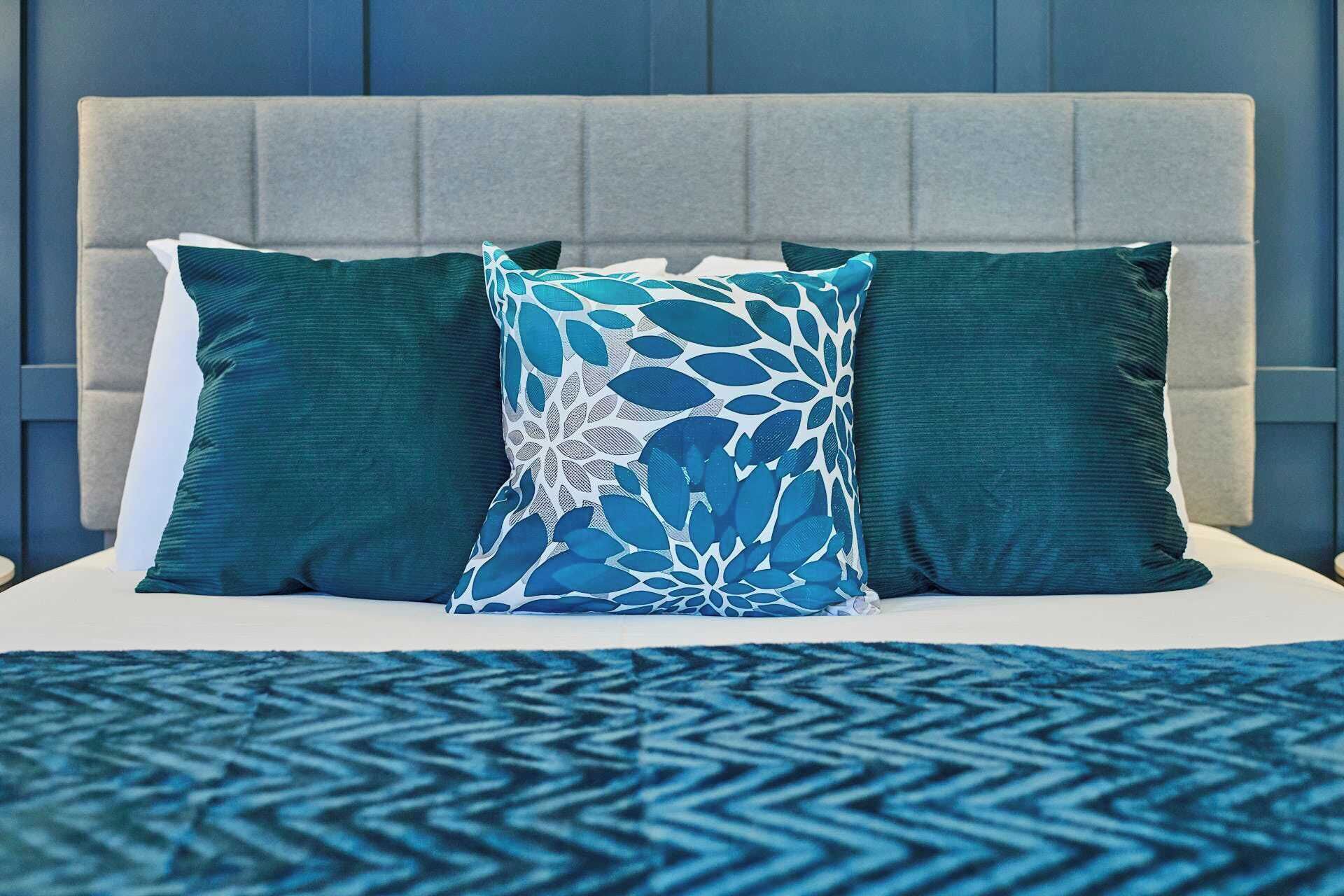 A bed with a grey upholstered headboard, two solid teal pillows, and a patterned pillow with a leaf design.