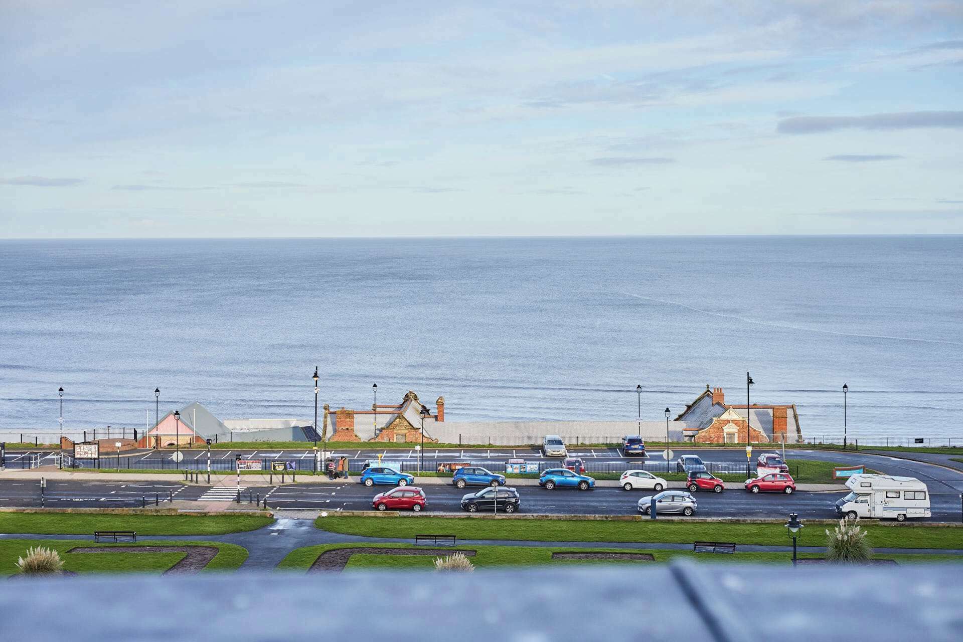 A coastal parking area with vehicles, a pedestrian walkway, and a view of the sea.