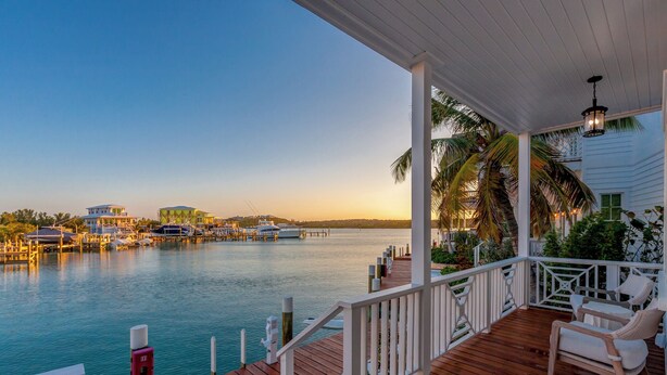 A wooden deck with white railings overlooking a marina with boats and buildings. - 1 Bedroom Villa - Sunset Point, Abaco -SeaScape Villa # 9 (Elbow cay)