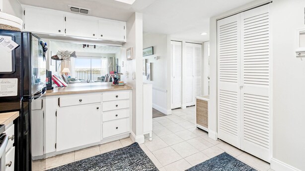 A modern kitchen with white cabinetry, a black refrigerator, and a wooden countertop. There are two white doors with louvered tops, a small window, and a tiled floor. - Charming Waterview Condo | 6 min wlk to beach! (Indian Shores)