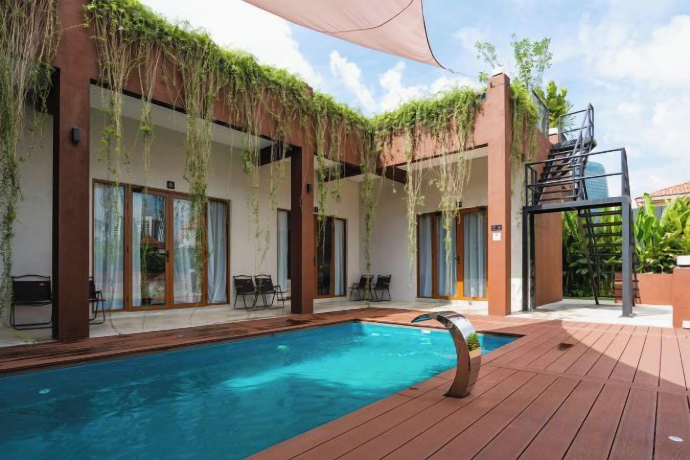 A swimming pool with a wooden deck, outdoor seating, and a covered area with plants.