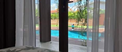 A view through a window with sheer curtains, revealing a swimming pool and greenery outside.